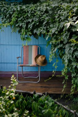 Tahta bir güvertede yeşil asma yapraklarıyla çevrili bir yaz şapkası ve katlanır bir sandalye. Kır evi estetiği. Şehir dışında bir tatil. Sıcak yaz günü.