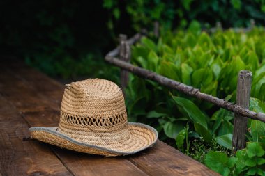 Bahçede yeşil yapraklarla çevrili ahşap bir güvertede eski bir hasır şapka. Bahçıvanlık, yaz, seyahat, tatil ya da dinlenme konsepti. Metnin için yer.