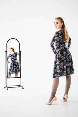 Güzel bir genç kadın model aynanın yanında duruyor ve beyaz bir arkaplanda kameraya bakıyor. Kameraya arkası dönük ve aynaya yansıyan elbise giymiş bir kızın stüdyo fotoğrafı..