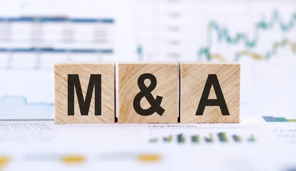 M and A or Merger and Acquisition made with wood building blocks on background from financial graphs and charts