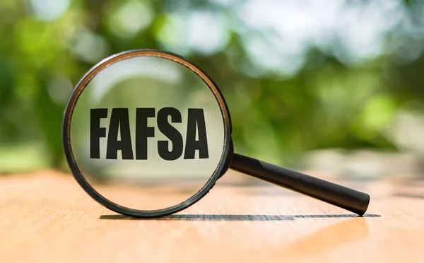 Federal Öğrenci Yardımı İçin Ücretsiz Başvuru - Ahşap masa ve yeşil arka planda büyüteç üzerine yazılmış FAFSA. Resmi kavra