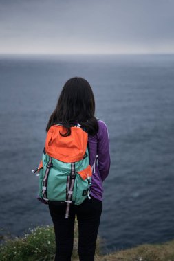 Okyanusa bakan düşünceli kadın. Deniz kenarında renkli bir sırt çantası taşıyan genç bir kadının arkası.
