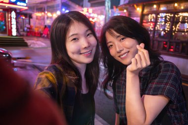 İki genç Asyalı Koreli bayan arkadaş cep telefonuyla kendi portresini çekiyorlar ve geceleri gülümseyerek poz veriyorlar.