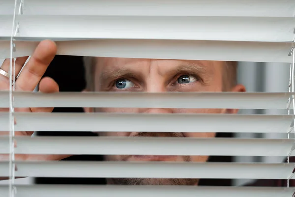 Man looking looks scared in window peeking out from blinds due to quarantine