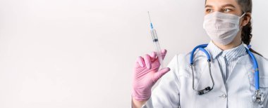 Beyaz önlüklü, tıbbi maskeli ve pembe steril eldivenli genç bir kadın doktor şırınga tutuyor. Şırıngaya odaklan. Aşılama kavramı.