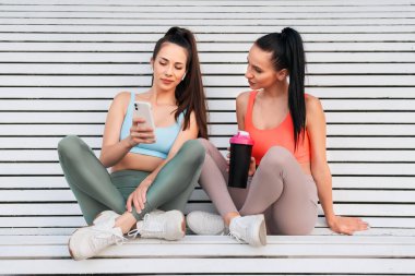 Genç kadınlar açık havada spor yaptıktan sonra rahatlarlar. Bir kız telefonunda bir şey gösteriyor.