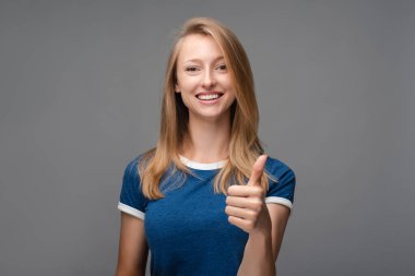Sarı düz saçlı neşeli kız, beyaz dişlerini gösteriyor, geniş gülümsemeli, kameraya mutlu bir ifadeyle bakıyor, iyi bir jest gösteriyor. İnsani duygular, yüz ifadesi kavramı.