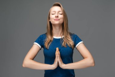 Genç sarışın kadın yoga yapıyor namaste 'de el ele tutuşuyor ve gözlerini kapalı tutuyor. Evde meditasyon yapıyor, huzur ve sevgi için dua ediyor, sakin ve huzurlu bir yüz ifadesi var.