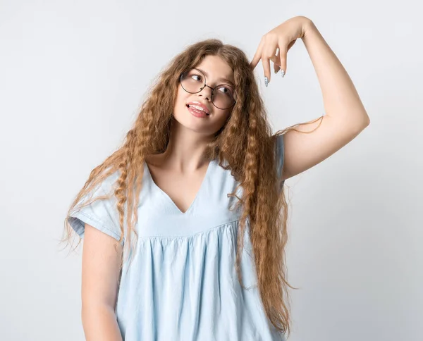 Photo of Pensive girl with curly long hair, and in round spectacles, looking up having pensive expression and holding her hand near head. Isolated over white background.