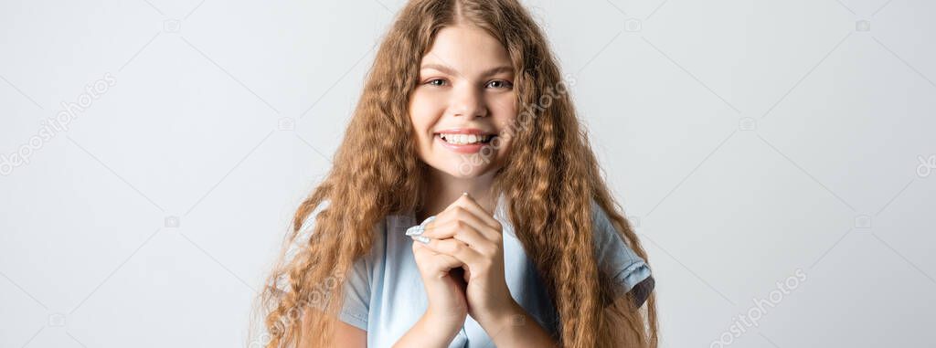 Photo of charismatic European young woman with curly long hair keeps hands together near chin, smiles gently, has cute expression, wears blue t-shirt.