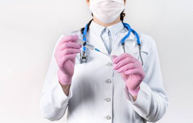Beyaz önlüklü, tıbbi maske takmış ve pembe steril eldivenli genç kadın doktor elinde şırınga tutuyor. Şırıngaya odaklan. Aşılama kavramı.