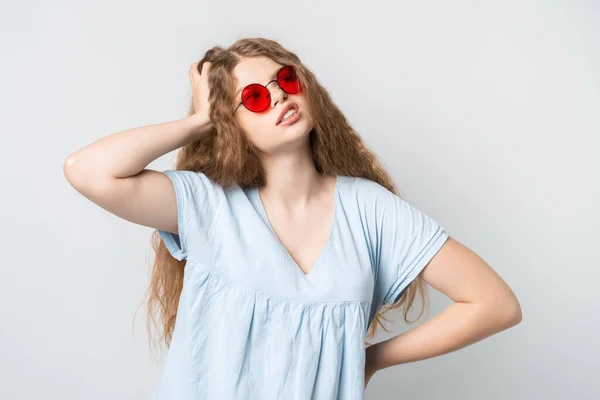 Photo of Pensive girl with curly long hair, and in round red spectacles, looking up having pensive expression and holding her hand near head. Isolated over white background.