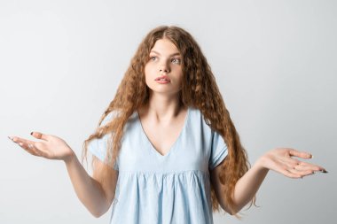 Ne oldu? Kafası karışmış, uzun kıvırcık saçlı güzel bir kızın portresi, ellerini yana açıyor, sırıtıyor, seçimler yaparken şüpheye düşüyor, gündelik tişört giyiyor. Beyaz arkaplanda izole.