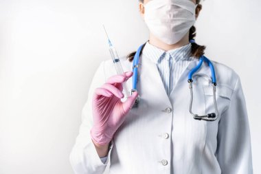 Beyaz önlüklü, tıbbi maske takmış ve pembe steril eldivenli genç kadın doktor elinde şırınga tutuyor. Şırıngaya odaklan. Aşılama kavramı.