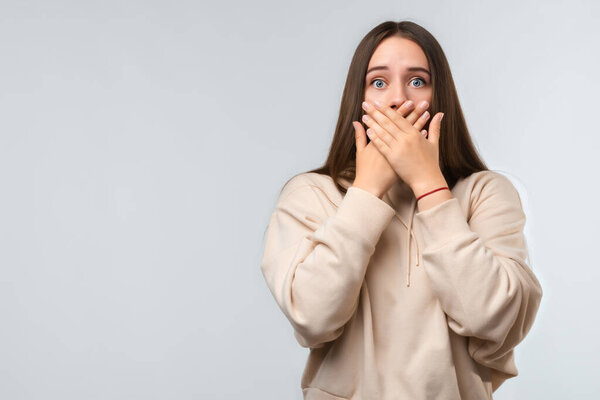 Шокированная молодая женщина с длинными каштановыми волосами, одетая в повседневную одежду, покрывающую рот руками на белом фоне. Понятие молчания