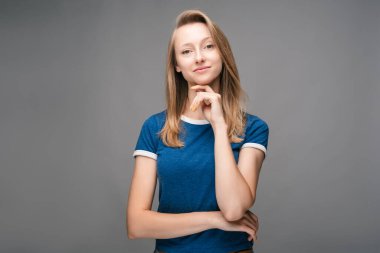 Sarışın, düz saçlı ve mavi tişörtlü düşünceli genç bir kadın. Gri arka planda izole edilmiş, el kaldırılmış bir şekilde kameraya bakıyor. Düşünce kavramı
