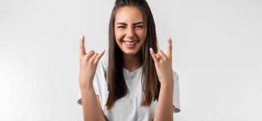 Uzun kestane rengi saçlı duygusal Avrupalı genç bir kadın Rock 'n Roll tabelasını yaptırıyor. Bu partiyi sallayacağımı, beyaz tişört giyeceğimi, beyaz arka planda boş bir alanda duracağımı ve kendime güvendiğimi söylüyor.