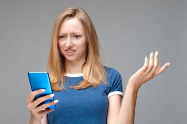 Sarı saçlı ve mavi tişörtlü rahatsız bir kız sorunlu bir yüz ifadesi var, akıllı telefon tutuyor, avuç içi kaldırıyor, sorunun ne olduğunu anlayamıyor, mesajın garip bir anlamı var, omuzlarını silkiyor.