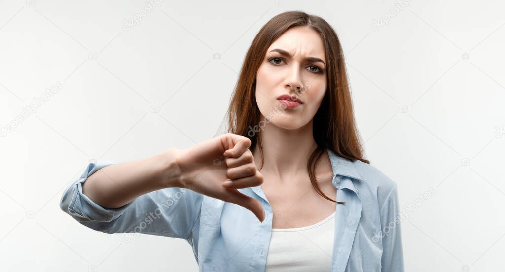 Beautiful caucasian girl with long chestnut hair, dressed in casual clothes, looking is upset at the camera, holding thumb down. Concept of dislike. Human emotions, facial expression concept