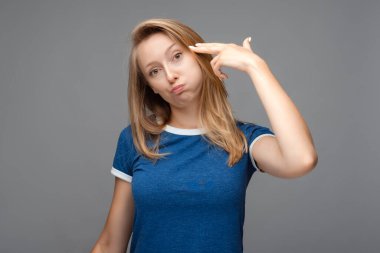 Komik sarışın kadın şakağına ateş ediyor, başını eğiyor, günlük mavi tişört giyiyor, intihar jesti yapıyor, gri arka planda izole edilmiş.