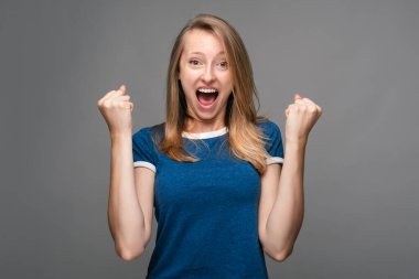 Başarılı, sarışın, düz saçlı, mavi tişörtlü, mutlu bir şekilde gülümseyen, harika haberleri kutlayan, kazanan bir jest yapan, gri arka planda duran genç bir kadın.