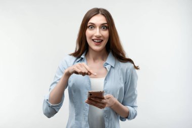 Uzun kestane rengi saçlı, sıradan kıyafetler giymiş beyaz arka planda akıllı telefon kullanan şaşırmış genç bir kadın. Yeni yararlı uygulama.