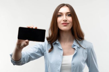 Uzun kestane rengi saçları olan, günlük giysiler giymiş, Akıllı Telefon Boş Ekran Gösteren, Beyaz Stüdyo Arkaplanı Üzerinde Mobil Uygulama Uygulamasına bakan genç bir kadın..