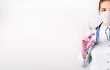 Beyaz önlüklü, tıbbi maske takmış ve pembe steril eldivenli genç kadın doktor elinde şırınga tutuyor. Şırıngaya odaklan. Aşılama kavramı.