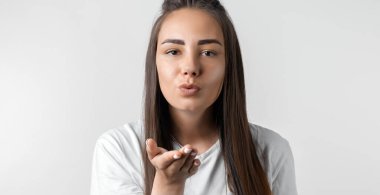 Uzun kestane rengi saçlı güzel Avrupalı genç bir kadın hava öpücüğü veriyor, avuç içi kadar üflüyor, erkeği öpmek istiyor. Sıradan kıyafetler giymiş, kameraya hava öpücüğü veren oyuncu bir kadın.