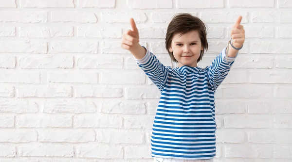 Approval gesture. Young boy 10-12 years old showing thumbs up against a white brick wall. Copy space for your text