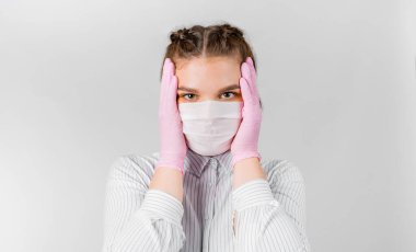 Beyaz gömlekli, tıbbi maskeli ve pembe steril eldivenli güzel bir kadın el ele tutuşuyor. Şok oldu ve başını tuttu. Kendini izole etme, karantina ve salgın hastalık kavramı.