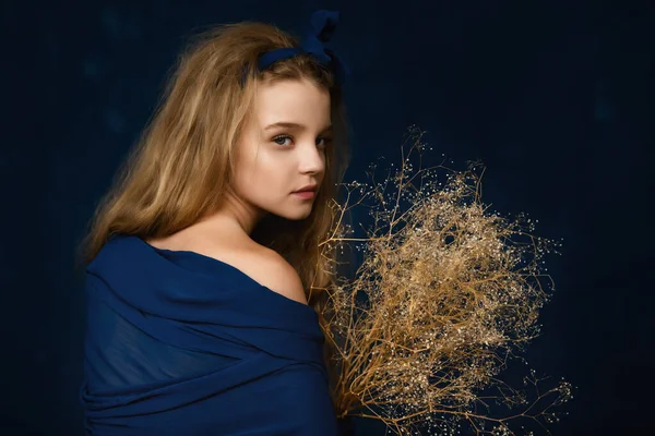Art Portrait of Beautiful young girl 10-12 years old with curly long hair, holding dried flowers in her hands on blue background. Fashion concept