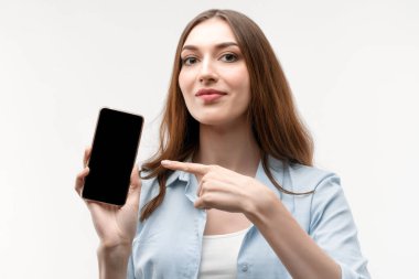 Uzun kestane rengi saçları olan, günlük giysiler giymiş, Akıllı Telefon Boş Ekran Gösteren, Beyaz Stüdyo Arkaplanı Üzerinde Mobil Uygulama Uygulamasına bakan genç bir kadın..