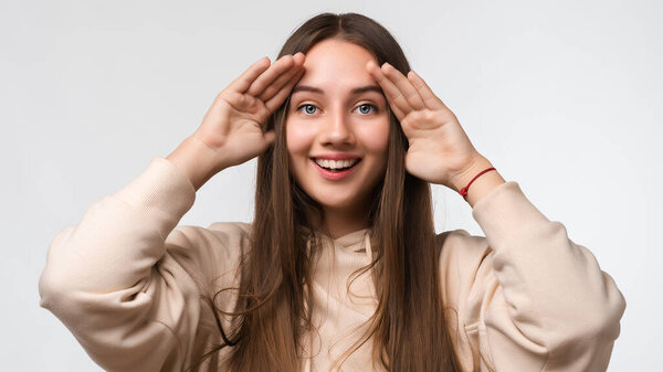 Close up studio portrait of a Young beautiful brunette woman looking through her fingers like binoculars, searching for something, wearing stylish beige hoodie, isolated on a white background
