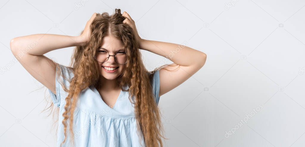 Oh NO. Photo of beautiful emotional girl with curly long hair and in round spectacles, looking at the camera, astonished, holding hands on head. Problems, surprise, fear, fobia concept