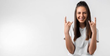 Uzun kestane rengi saçlı duygusal Avrupalı genç bir kadın Rock 'n Roll tabelasını yaptırıyor. Bu partiyi sallayacağımı, beyaz tişört giyeceğimi, beyaz arka planda boş bir alanda duracağımı ve kendime güvendiğimi söylüyor.