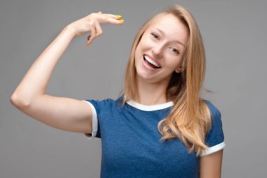 İyi ki sarışın bir kadın şakakta ateş ediyor, başını eğiyor, ağzını geniş bir şekilde açık tutuyor, günlük mavi tişört giyiyor, intihar jesti yapıyor, gri arka planda izole edilmiş.