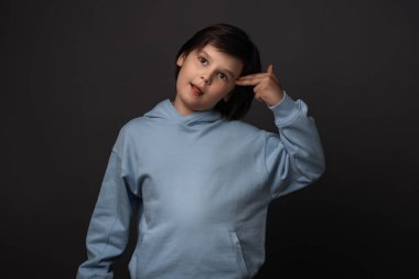 Kafası karışmış 10-12 yaşlarında, gündelik giysiler giymiş, kendini vuracakmış gibi parmağıyla başını işaret eden bir çocuk. Stüdyo fotoğrafı, gri arka plan. İnsani duygular, yüz ifadesi kavramı