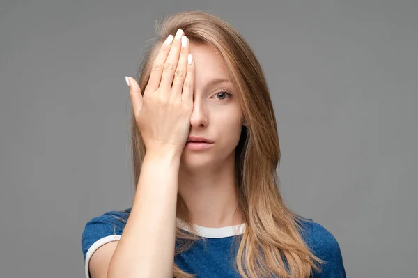 Yorgun, sarışın, yüzü örtülü, yorgun, iyi dinlenmesi gereken, mavi tişörtlü, gri arka planda izole genç bir kadın fotoğrafı. İnsani duygular, yüz ifadesi kavramı.