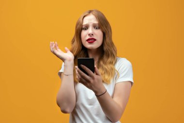 Uzun dalgalı sarı saçlı, beyaz tişörtlü, sarı arka planda akıllı telefon kullanan genç bir kadın. Yeni yararlı uygulama, hızlı limitsiz internet kullanıyor, online alışveriş yapıyor