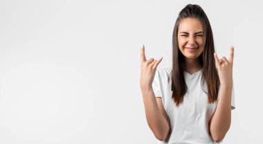 Uzun kestane rengi saçlı duygusal Avrupalı genç bir kadın Rock 'n Roll tabelasını yaptırıyor. Bu partiyi sallayacağımı, beyaz tişört giyeceğimi, beyaz arka planda boş bir alanda duracağımı ve kendime güvendiğimi söylüyor.