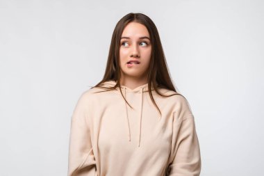 Düşünceli bir kadının fotoğrafı alt dudağını ısırıyor, rüya gibi bir ifadeyle bakıyor, şık bir bej kapüşon giyiyor, beyaz duvarın karşısında duruyor.