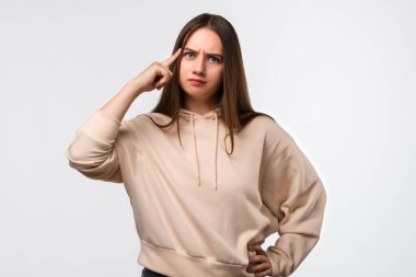 Uzun kestane rengi uzun saçlı, başı parmağıyla gösterilen, şık bej kapüşonlu genç bir kadının stüdyo fotoğrafı. Düşünce anlayışı. Akıllı ol ve düşün..