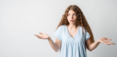 Ne oldu? Kafası karışmış, uzun kıvırcık saçlı güzel bir kızın portresi, ellerini yana açıyor, sırıtıyor, seçimler yaparken şüpheye düşüyor, gündelik tişört giyiyor. Beyaz arkaplanda izole.
