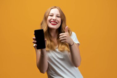 Uzun dalgalı sarı saçlı, beyaz tişörtlü, Smartphone Boş Ekran Gösteren, Sarı Stüdyo Arkaplan Üzerinde Mobil Uygulama Kamerasına bakan genç bir kadın.