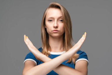 Sarışın, düz saçlı, mavi tişörtlü, çapraz elli, dur, bitir, reddedilmenin sembolünü gösteren, şiddete izin vermeyen, vücut diline izin vermeyen ciddi bir kız..