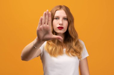 Dalgalı kızıl saçlı, ciddi ve sert görünümlü, beyaz tişört giyen, avuç içi açık duran genç ve güzel bir kadın. Sarı arkaplan üzerinde izole edilmiş