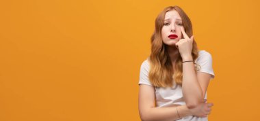 Dalgalı kızıl saçlı, beyaz tişörtlü, gözü işaret eden güzel bir kız jestini izliyor, şüpheli bir ifade. Sarı arka planda izole edilmiş. Metnin için boşluk kopyala