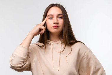 Uzun kestane rengi uzun saçlı, başı parmağıyla gösterilen, şık bej kapüşonlu genç bir kadının stüdyo fotoğrafı. Düşünce anlayışı. Akıllı ol ve düşün..