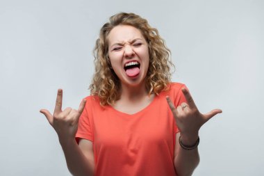 Heyecanlı, kıvırcık saçlı, sıradan tişörtlü, gülen ve parmaklarıyla boynuz hareketi yapan sarışın kızın görüntüsü şöyle diyor: Bu partiyi sallayacağım. İnsan duyguları konsepti. Stüdyo çekimi, beyaz arkaplan